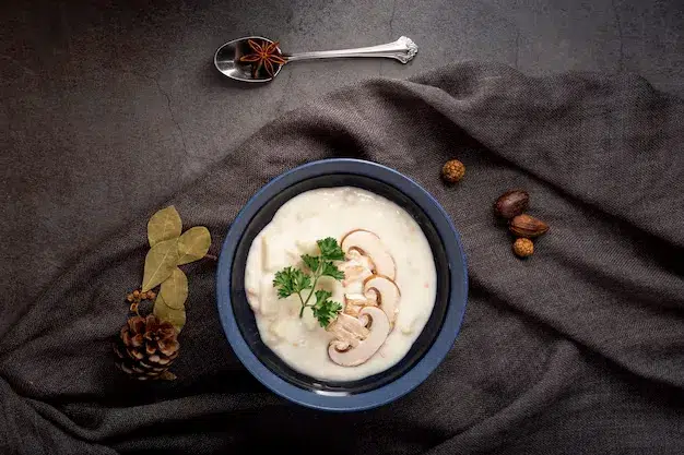 A bowl of creamy rice dish garnished with fresh parsley and thinly sliced mushrooms, placed on a textured dark background with decorative elements like pine cones, bay leaves, and spices It Shows Helixx Foods quality
