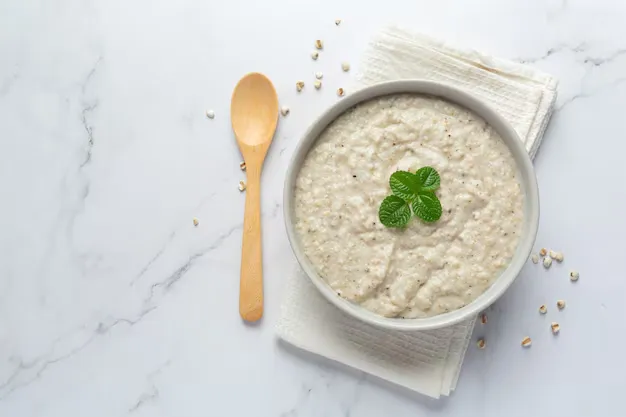 A simple bowl of creamy rice topped with a sprig of mint, placed on a white napkin with a wooden spoon and scattered grains on a marble background.