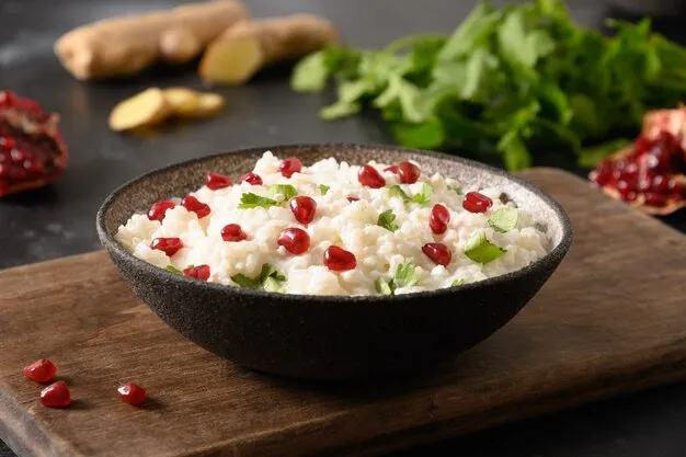 A black bowl of creamy rice garnished with fresh pomegranate seeds and coriander, placed on a wooden board with ingredients like ginger and leafy greens in the background.